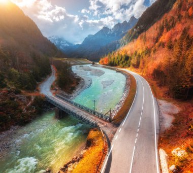 Sonbaharda günbatımında dönen dağ yolunun hava manzarası. Alpler, İtalya. Alp yolu, köprü, nehir, karlı kayalar, renkli ağaçlar, kırmızı yapraklar, göl, bulutlu gökyüzü. Sonbaharda dağın tepesindeki yol manzarası