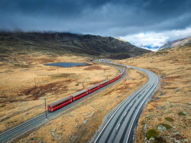 Alp dağlarındaki modern kırmızı trenin havadan görünüşü, yol, turuncu orman, sonbaharda günbatımında bulutlu gökyüzü. Bernina Express, İsviçre. Alplerdeki trenin en iyi insansız hava aracı görüntüsü, sonbaharda demiryolunu saran...