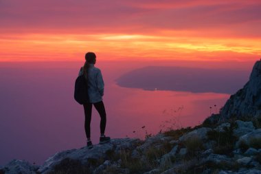 İtalya 'da gün batımında kayalık dağın zirvesinde duran sırt çantalı kadın yürüyüşçü dramatik pembe gökyüzü ile denize bakıyor. Açık hava macerası. Alacakaranlıkta dağda sportif bir kız