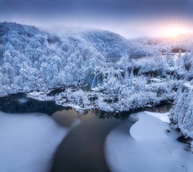 Aerial drone view of frozen lakes and waterfalls surrounded by snowy forest at sunrise in Plitvice Lakes National Park, Croatia. Winter landscape with icy cascades and scenic European nature. Top view