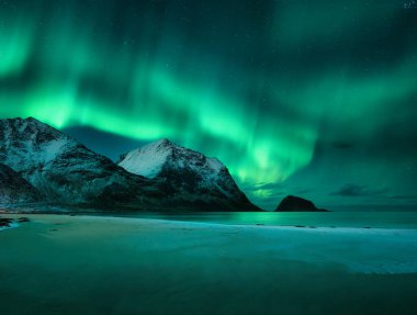 Aurora Borealis, gece gökyüzünü karlı dağların üzerinde yeşille boyuyor ve kumlu bir plaj ışığı yansıtıyor. Norveç, Lofoten adalarında kış harikalar diyarı. Kuzey ışıkları, yıldızlı gökyüzü, deniz, kaya