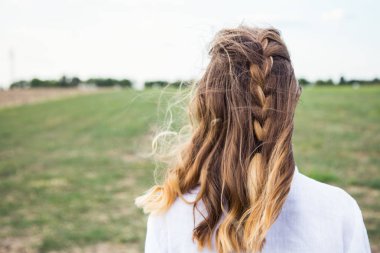 Portrait of young blonde from behind with carelessly braided pigtail and flying hair in wind in field. Concept of harmony and calmness. clipart