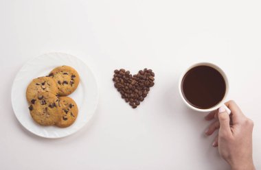 Fincan kahve, çikolata kurabiye ve beyaz zemin üzerine kavrulmuş fasulye kalp tutan el. Tonda düz lay.