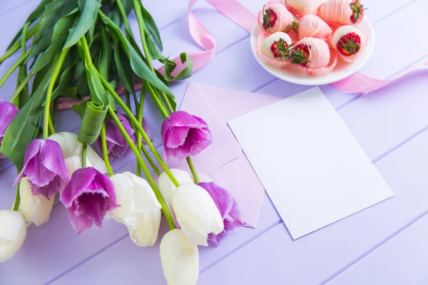 Clear sheet of paper with envelope, strawberry in chocolate, bouquet of white and purple tulips and pink ribbon on lilac table. Holiday greetings for woman or mother. Spring decoration.