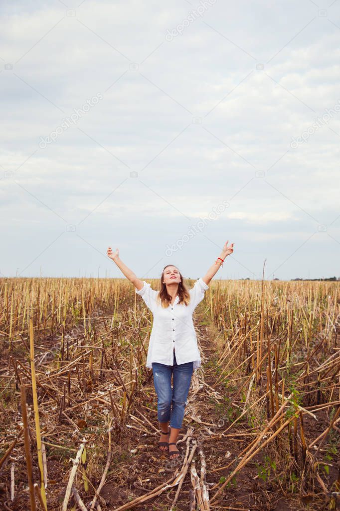 Young blond girl in casual clothes with arms raised enjoys her walking in field. Happy woman relaxing on countryside. Concept of freedom and wellness.