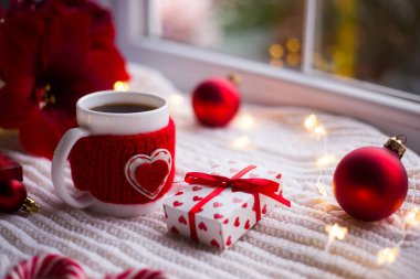 Örme rahat Kupası Kahve ve hediye kutusu üzerinde beyaz ekose kış dekorasyonu ile çevrili. Nergis zambağı, kırmızı Noel topları, hediye kutusu ve gün ışığından yararlanma penceresinde yakınındaki karamel köpekler. Yeni yıl tatil kompozisyon. Sevgililer günü kartı.