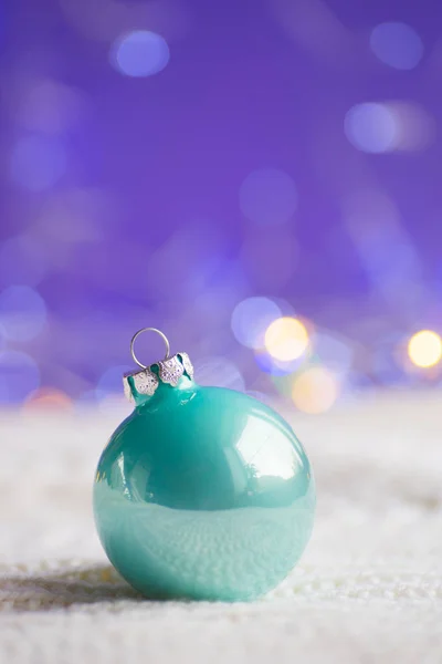 Pastel mint green Christmas ball on white knitted fabric on purple background with warm bokeh. New year card. Xmas decoration.