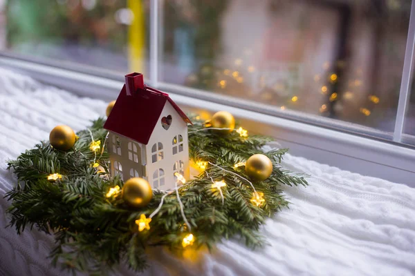 Toy house with hole in form of heart near fir wreath decorated with golden Christmas balls and coiled with glowing garland with warm light on white knitted plaid. New Year home decoration