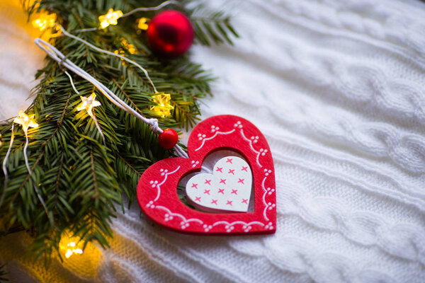 Wooden heart near fir wreath decorated with Christmas balls and coiled with glowing garland with warm light on white knitted plaid. New Year home decoration. St. Valentine Day card.