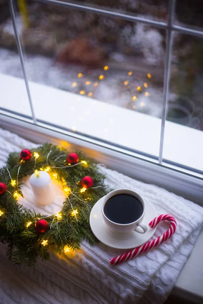 White cup of coffee and caramel cane near fir wreath decorated with red Christmas balls, burning candle and coiled with glowing garland with warm light near window. New Year home atmosphere.