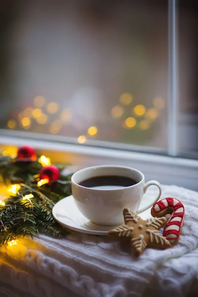 White cup of coffee and Christmas gingerbread near fir wreath decorated with red balls, burning candle and coiled with glowing garland with warm light near window. New Year evening at home.