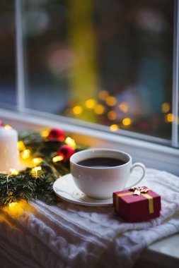 Kahve ve hediye kutusu köknar çelenk yakınındaki beyaz fincan kırmızı Noel topları, yanan mum ile dekore edilmiş ve pencere yakınındaki sıcak ışığıyla parlayan garland ile kıvrılmış. Yeni yıl eve atmosfer
