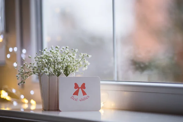 Greeting card with best wishes and bouquet of beautiful white gypsophila near window in the daylight, bokeh lights on background. Tender romantic banner