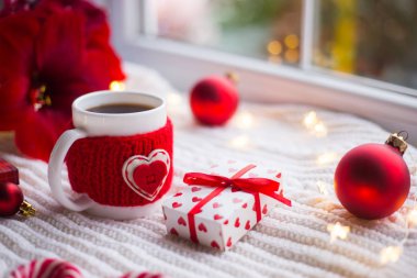 Örme rahat fincan çay kalp ve kış dekorasyonu ile çevrili beyaz örme şal üzerinde romantik hediye kutusu ile. Nergis zambağı, Noel topları ve garland ışıklar Günışığı penceresinde yakınındaki. St Sevgililer günü kartı.