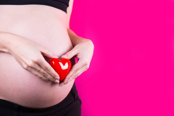 孕妇是拿着红色塑料心脏在她的手在肚子附近在粉红色的背景 母亲的爱和幸福的期待的概念的婴儿 带复制空间的工作室拍摄 — 图库照片