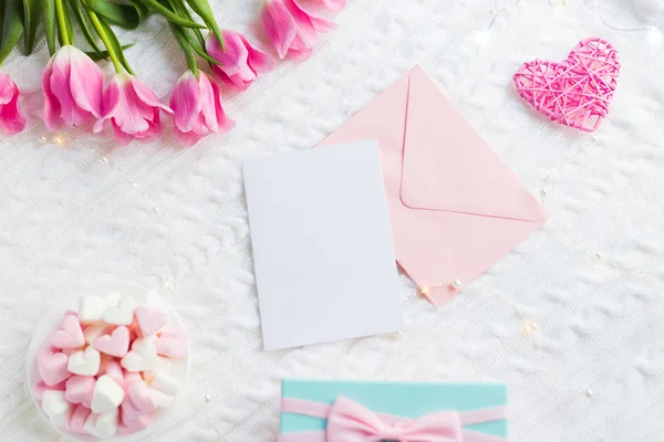 Clear paper and envelope with gift box, pink tulips, heart and marshmallow on white knitted plaid, top view. Card with copy space. Spring romantic composition for text, tender flat lay.