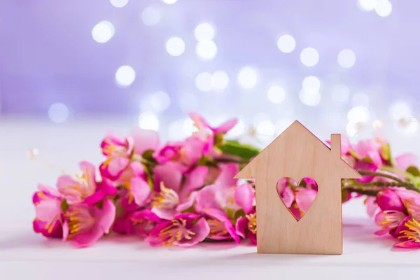 Closeup wooden house with hole in form of heart with tender pink cherry flowers on blurred lilac background with bokeh. Spring romantic composition with copy space