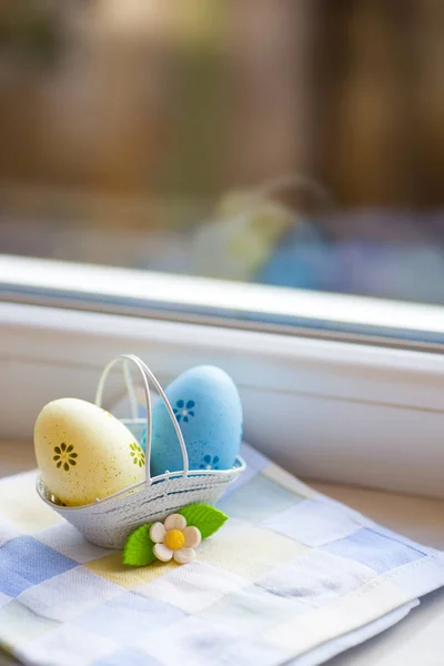 Coloridos huevos de Pascua azules y amarillos en cesta con flores decorativas cerca de la ventana a la luz del día . — Foto de Stock