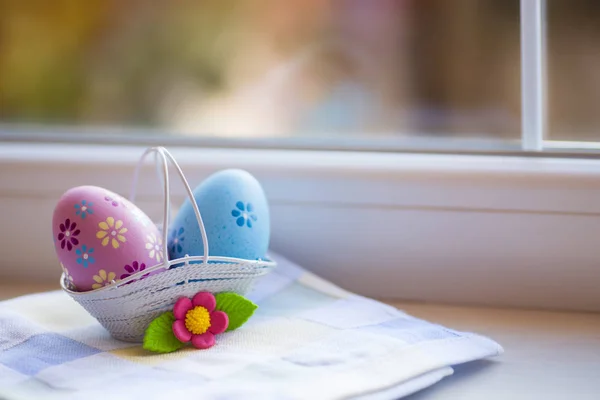 Colorate uova di Pasqua rosa e blu nel cestino con fiore decorativo vicino alla finestra alla luce del giorno . — Foto Stock