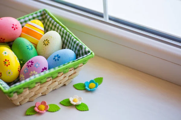 Variety of decorated colorful Easter eggs in checkered basket with spring flowers near window in daylight. — Stock Photo, Image