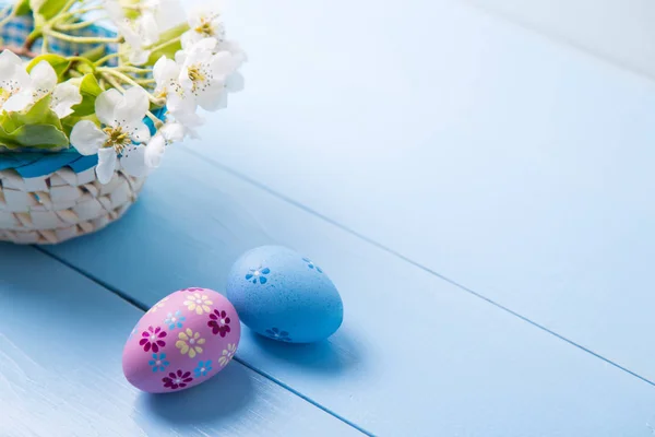 Two painted blue and purple Easter eggs near basket with white spring flowering branch on light blue background — Stock Photo, Image