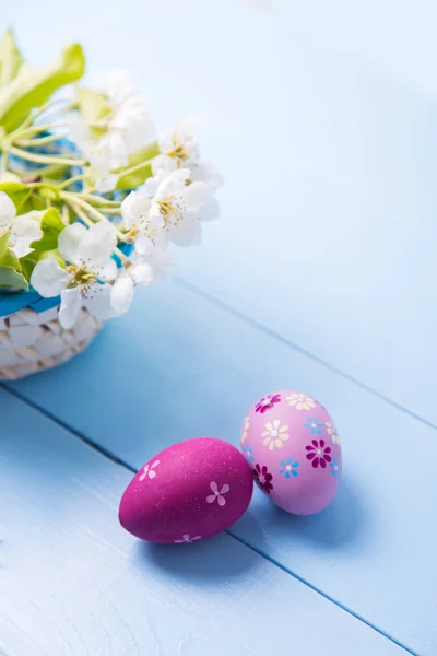 Dos huevos de Pascua pintados de color rosa y púrpura cerca de la cesta con rama floreciente de primavera blanca sobre fondo azul claro — Foto de Stock