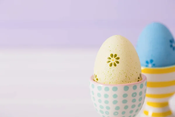 Nahaufnahme zwei bunt bemalte Ostereier in lebendigen modernen Eierständen auf pastellfliederfarbenem Hintergrund. — Stockfoto