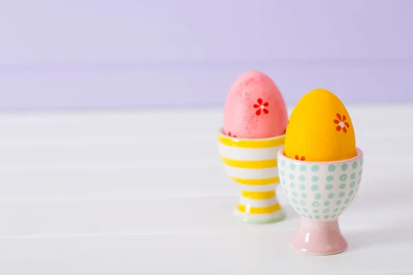 Nahaufnahme zwei bunt bemalte Ostereier in lebendigen modernen Eierständen auf pastellfliederfarbenem Hintergrund. — Stockfoto