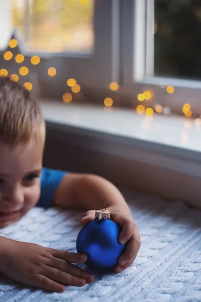Happy child boy holding blue Christmas ball near window indoor with warm garland lights on blurred background.