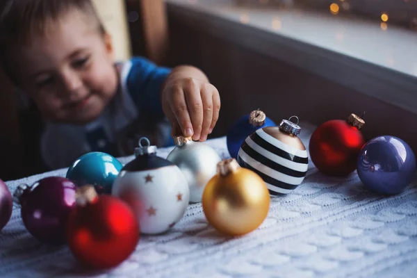 Happy child boy choosing Christmas ball near window indoor with warm garland lights on blurred background.