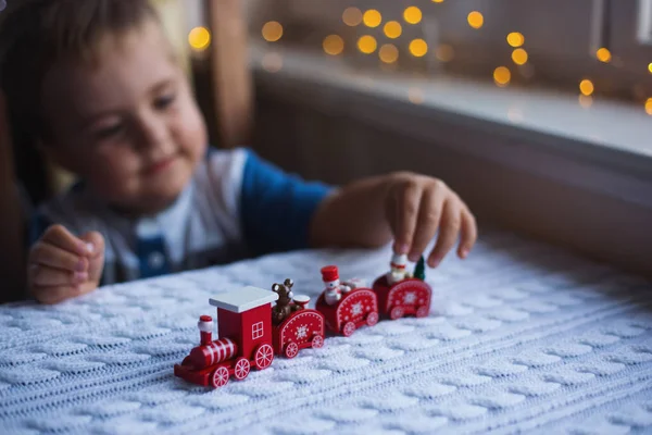 Niño jugando con juguete rojo tren de Navidad en punto blanco a cuadros a la luz del día con guirnalda cálida fondo bokeh — Foto de Stock