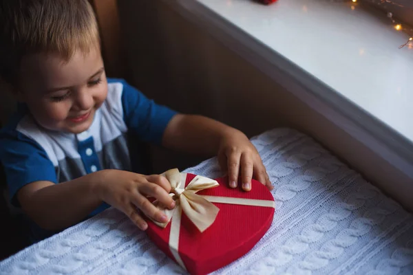 Niño feliz sosteniendo caja de regalo en forma de corazón rojo con arco cerca de la ventana a la luz del día . — Foto de Stock