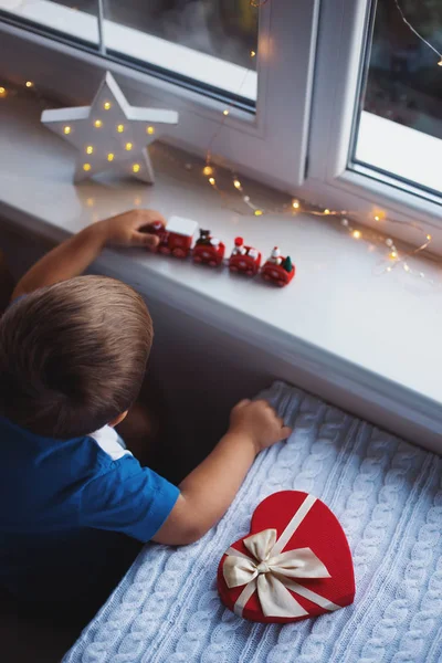 Niño jugando juguete tren rojo cerca de la ventana a la luz del día. Caja de regalo roja en forma de corazón con lazo en la mesa. Un regalo de San Valentín. Sorpresa para la persona amada y querida . — Foto de Stock