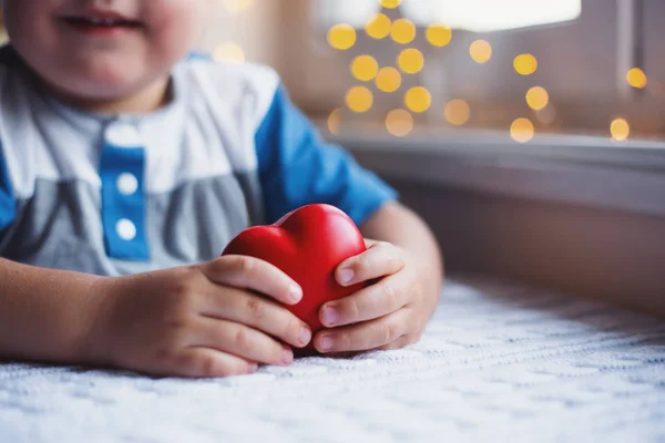 Niño sonriente sosteniendo el corazón de juguete rojo en las manos cerca de la ventana a la luz del día con acogedor bokeh en el fondo. El concepto de amor por los niños y el cuidado cardiológico . — Foto de Stock