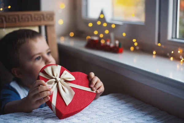 Niño niño sosteniendo caja de regalo en forma de corazón rojo con arco cerca de la ventana a la luz del día con luces bokeh cálidas en el fondo. regalo de San Valentín . — Foto de Stock