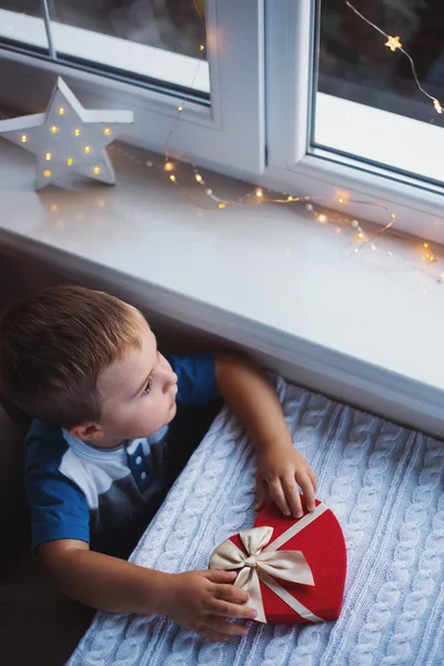 Niño pequeño sosteniendo caja de regalo en forma de corazón rojo con arco cerca de la ventana a la luz del día . — Foto de Stock