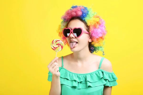 girl in clown wig showing tongue out and holding lollipop on yellow background