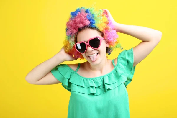 girl in clown wig and sunglasses showing tongue out on yellow background