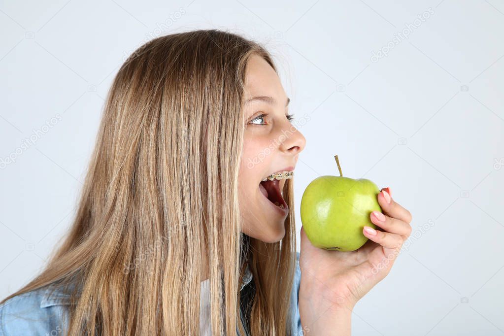 Young smiling girl with dental braces biting green apple on grey background
