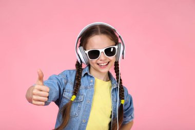 Kulaklıklar müzik dinlemek ve başparmak, pembe arka plan gösterilmesini güneş gözlüklü genç kız