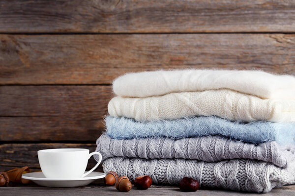 Stack of knitted sweaters with cup of tea, acorns and chestnuts on wooden background