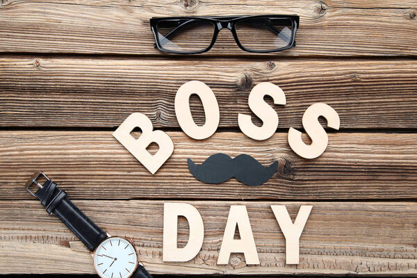 Inscription Boss Day with wrist watch and glasses on wooden table
