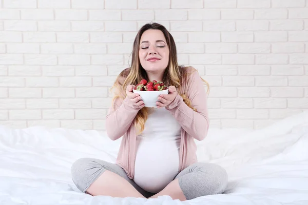 ベッドの上に座ってボウルにイチゴと美しい妊娠中の女性 — ストック写真