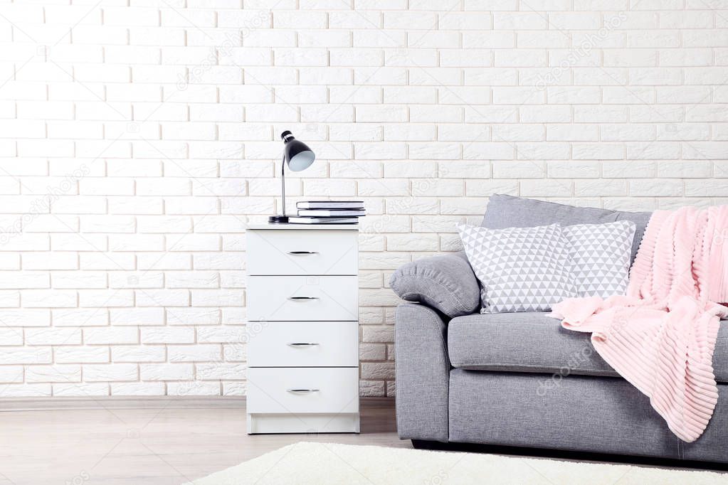 White bedside table near grey sofa with pillows and plaid on brick wall background