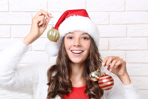 レンガ壁の背景にクリスマスのつまらないものを保持しているサンタ帽子で美しい少女 — ストック写真