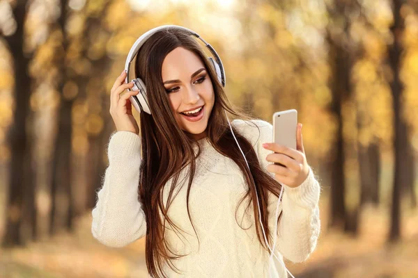 美丽的妇女与耳机和智能手机在秋天公园 — 图库照片