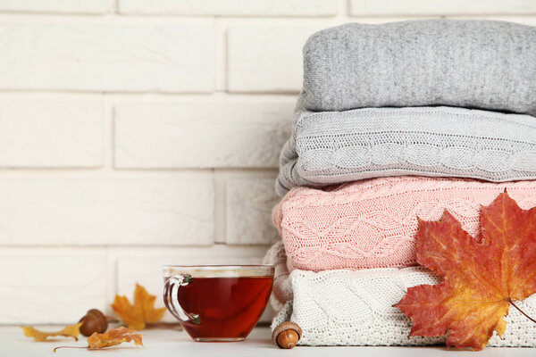Stack of knitted sweaters with cup of tea and autumn leaves on brick wall background
