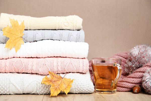 Stack of knitted sweaters with cup of tea and autumn leaves on wooden table