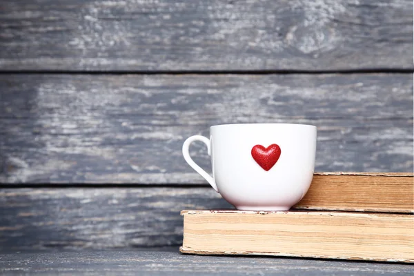 White cup with red heart and old books on grey wooden table