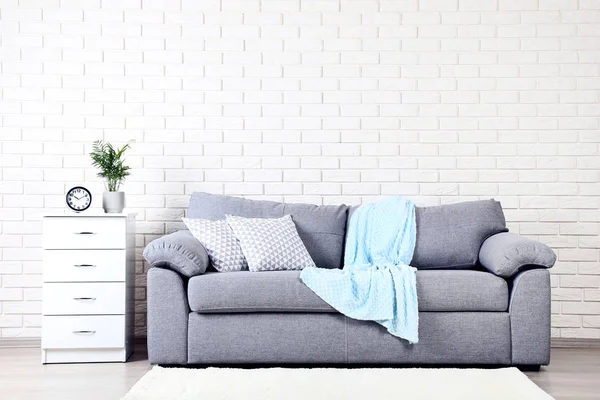 枕およびレンガ壁の背景に格子縞が付いている灰色ソファーの近く白いベッドサイド テーブル — ストック写真